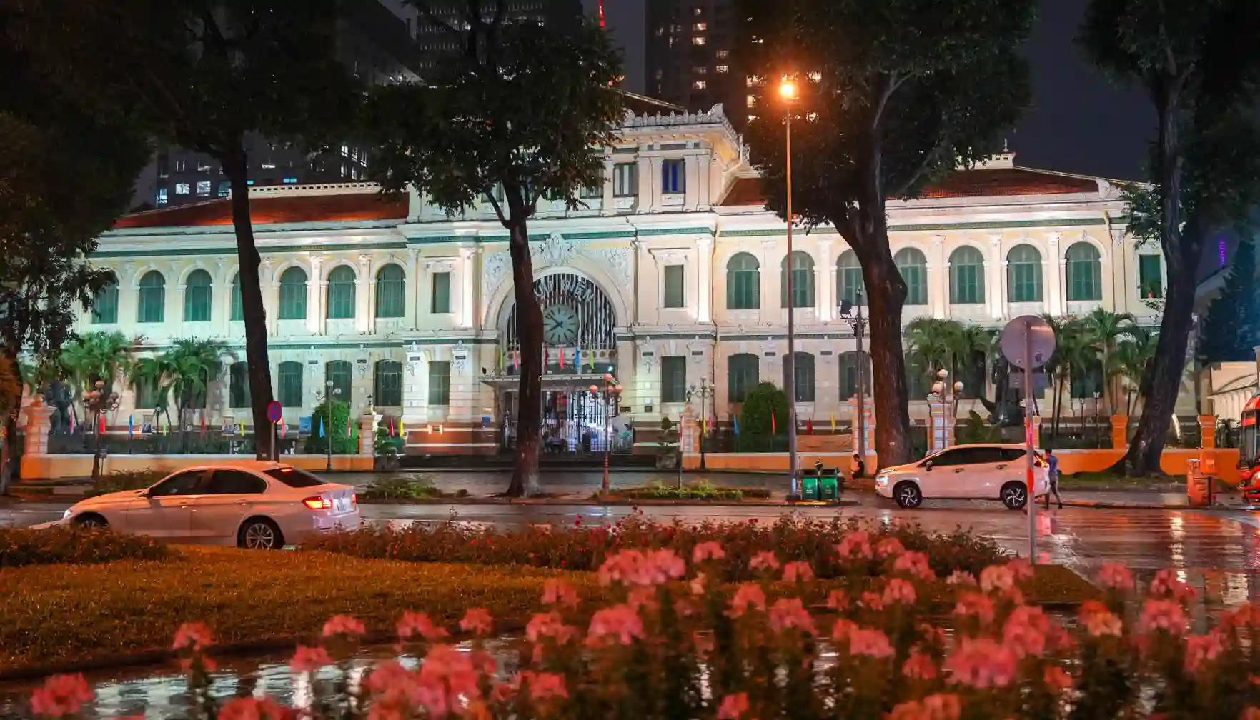 Saigon Central Post Office illuminated at night after rain, with French colonial facade, clock, and pink flowers in District 1, Ho Chi Minh City