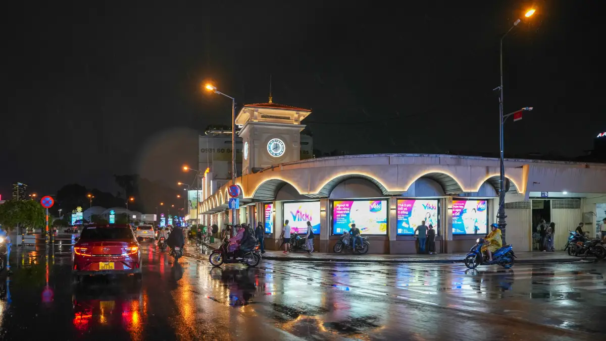 Ben Thanh Market at night after rain, with the illuminated clock tower and motorbikes on wet streets in District 1, Ho Chi Minh City