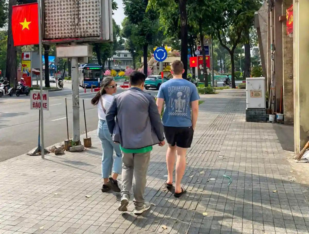 Travellers walking with their local guide along a tree-lined sidewalk in District 1, Ho Chi Minh City, past a Vietnamese flag and a roundabout