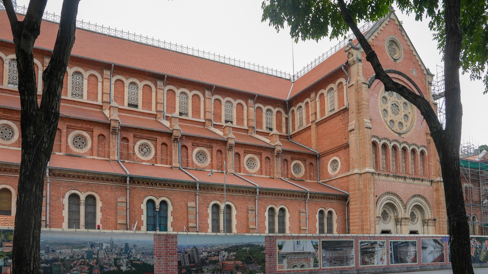 Red-brick side facade of Notre Dame Cathedral Saigon framed by mature trees, showing the rhythmic arched windows and rose details typical of French colonial Romanesque-Gothic architecture in District 1