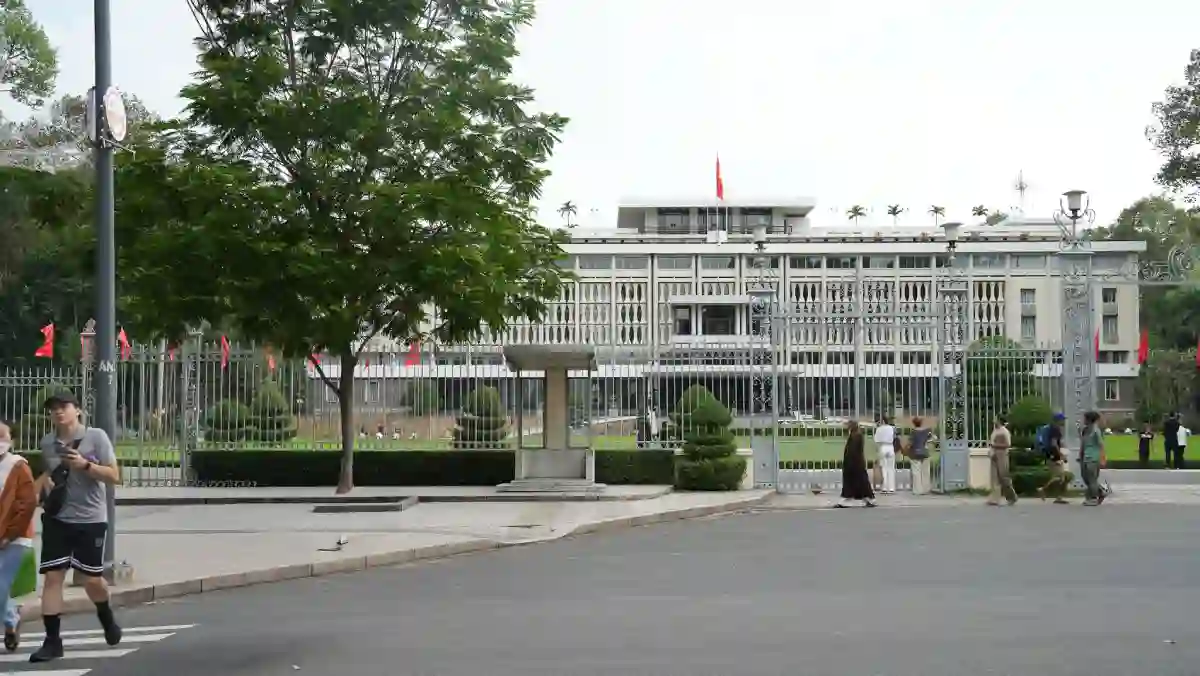 Independence Palace seen from the main gate on Nam Ky Khoi Nghia Street, with visitors walking past the white wrought-iron fence in District 1, Ho Chi Minh City
