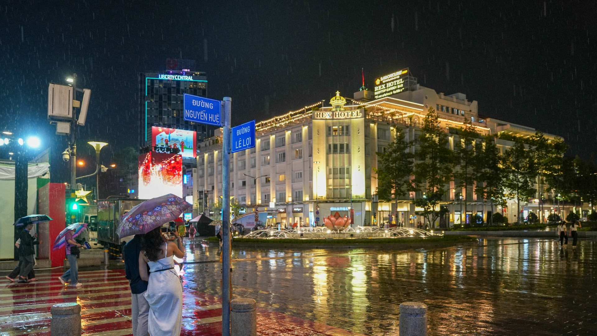 Blue Nguyen Hue and Le Loi street signs in the rain at night, with the illuminated Rex Hotel and the lotus fountain in the background, District 1, Ho Chi Minh City