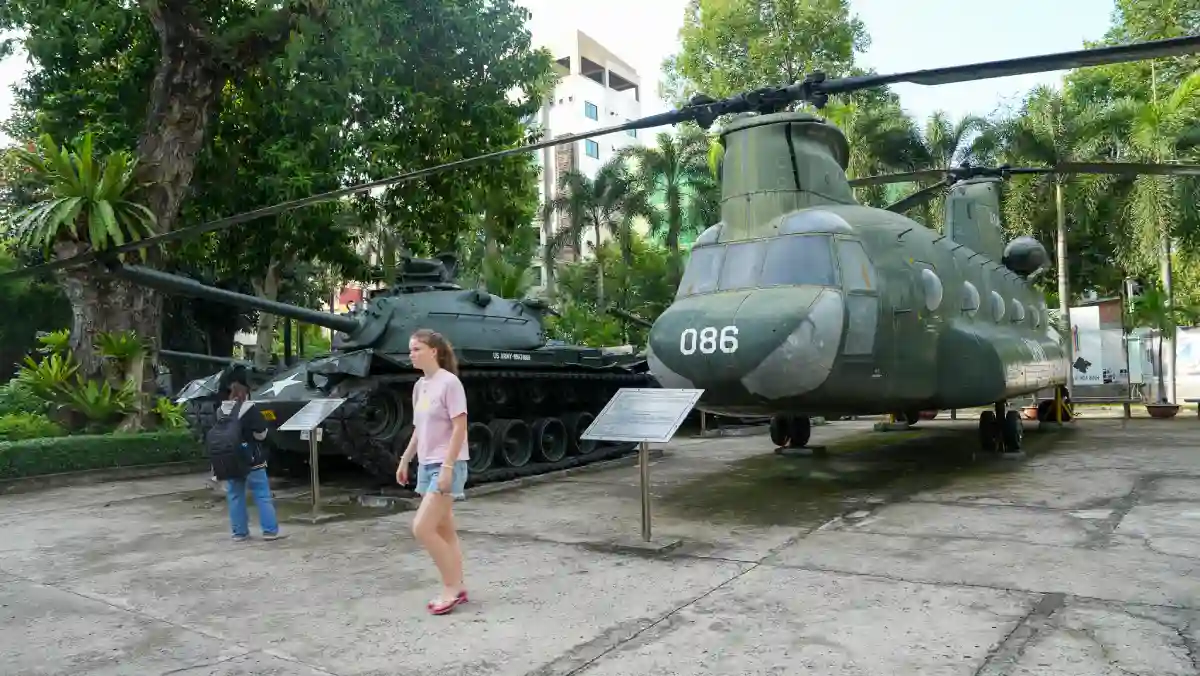 A CH-47 Chinook helicopter displayed beside a US Army tank in the outdoor courtyard of the War Remnants Museum, with modern Ho Chi Minh City buildings rising in the background