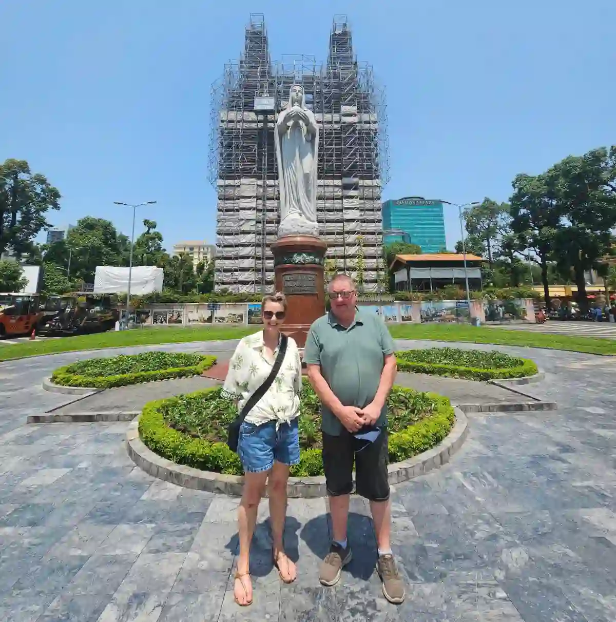 Two travellers in front of Notre Dame Cathedral Saigon during the ongoing restoration, with scaffolding around the bell towers and the Virgin Mary statue in District 1, Ho Chi Minh City