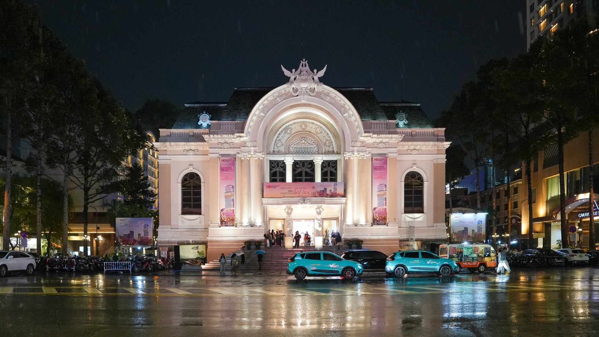 The illuminated Saigon Opera House at night in light rain, with Beaux-Arts façade glowing pink-cream above Lam Son Square in District 1, Ho Chi Minh City