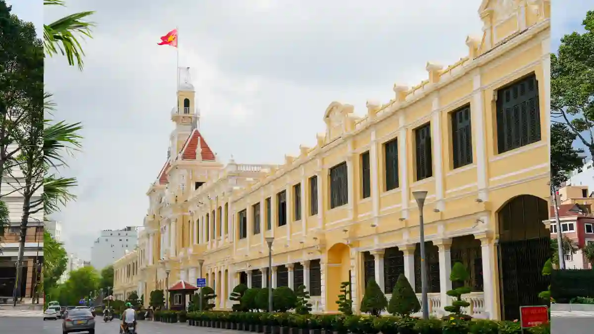 Ho Chi Minh City Hall — yellow colonial façade and clock tower with the Vietnamese flag, seen from the side along Le Thanh Ton Street in District 1, Saigon
