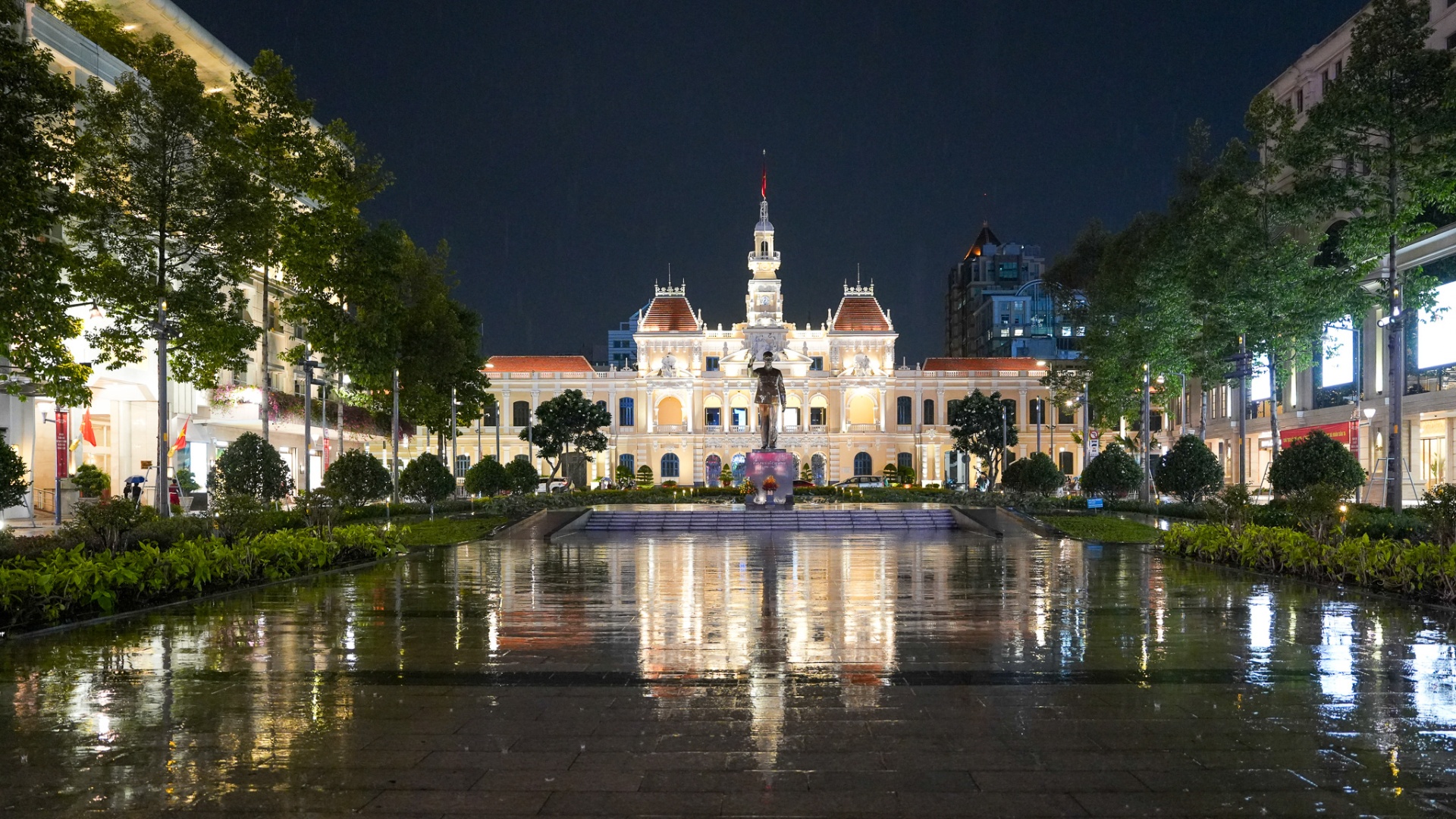 Ho Chi Minh City Hall illuminated at night after rain, reflected across the wet granite plaza of Nguyen Hue, with the Uncle Ho statue at its centre, District 1, Saigon