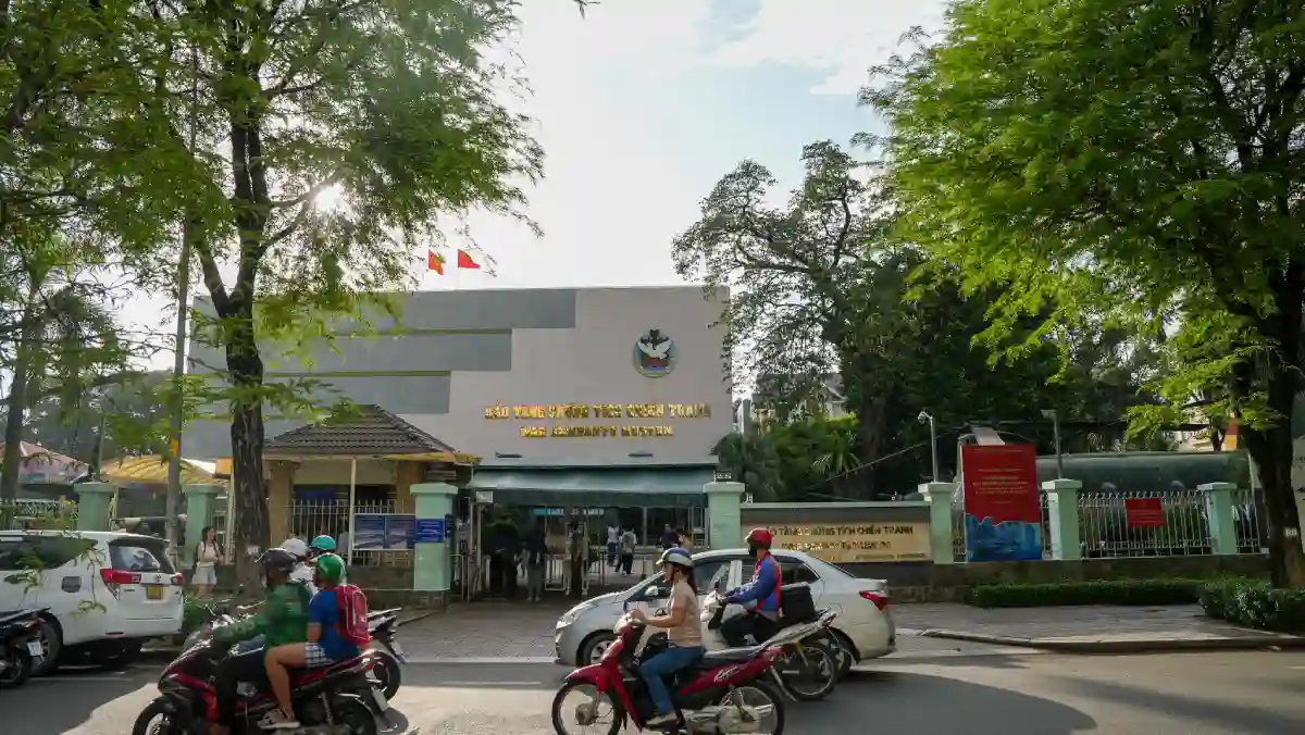 Main entrance of the War Remnants Museum on Vo Van Tan Street, District 3, Ho Chi Minh City, framed by tropical trees with motorbikes passing in the foreground