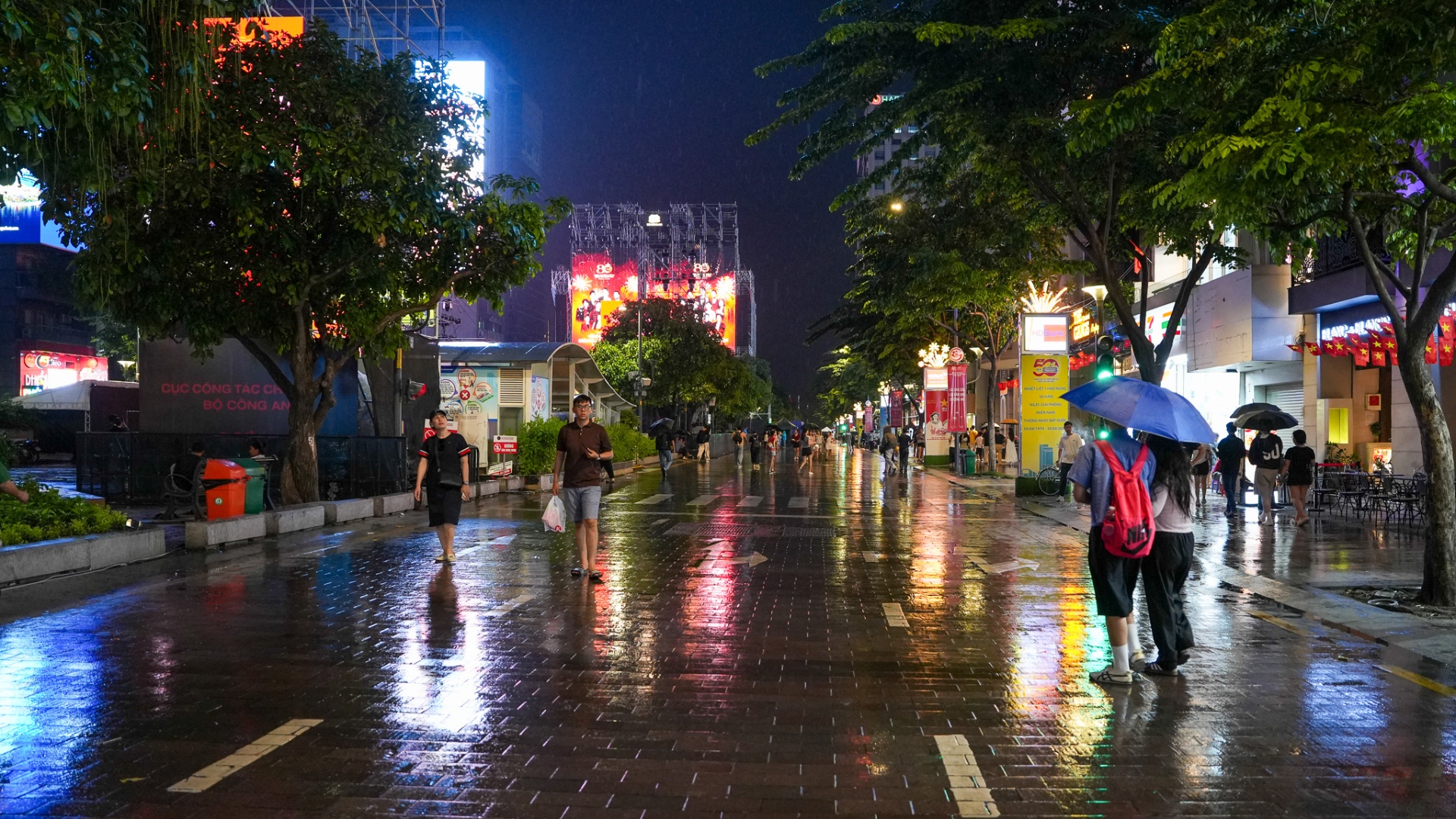 Rain-soaked Nguyen Hue Walking Street at night with pedestrians under umbrellas and a large LED stage glowing in the background, District 1, Saigon