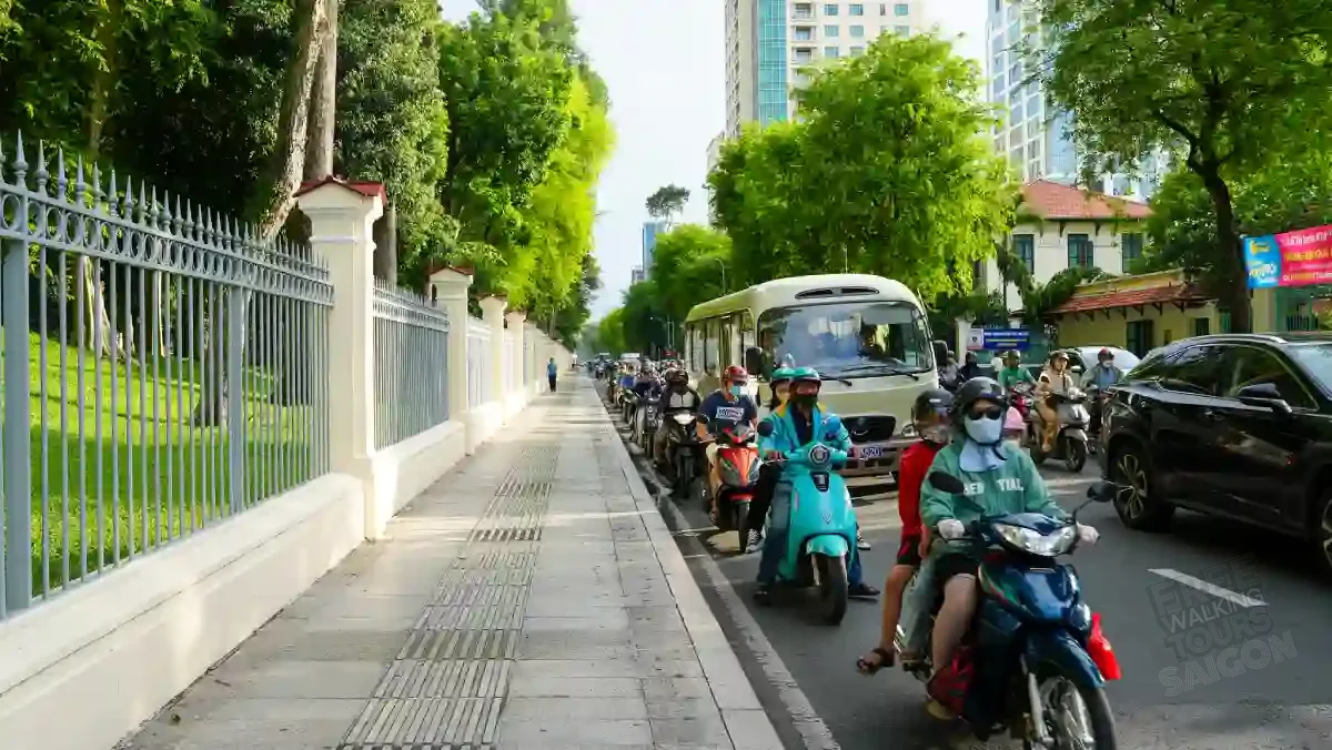 A tree-lined sidewalk in District 1, Ho Chi Minh City beside a row of motorbikes queueing along the street on a sunny afternoon