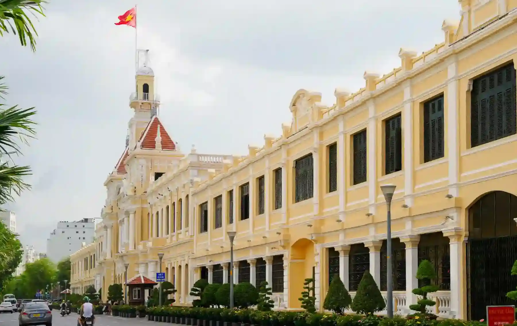 Ho Chi Minh City Hall — yellow French colonial façade and clock tower with the Vietnamese flag, seen along Le Thanh Ton Street in District 1, Saigon