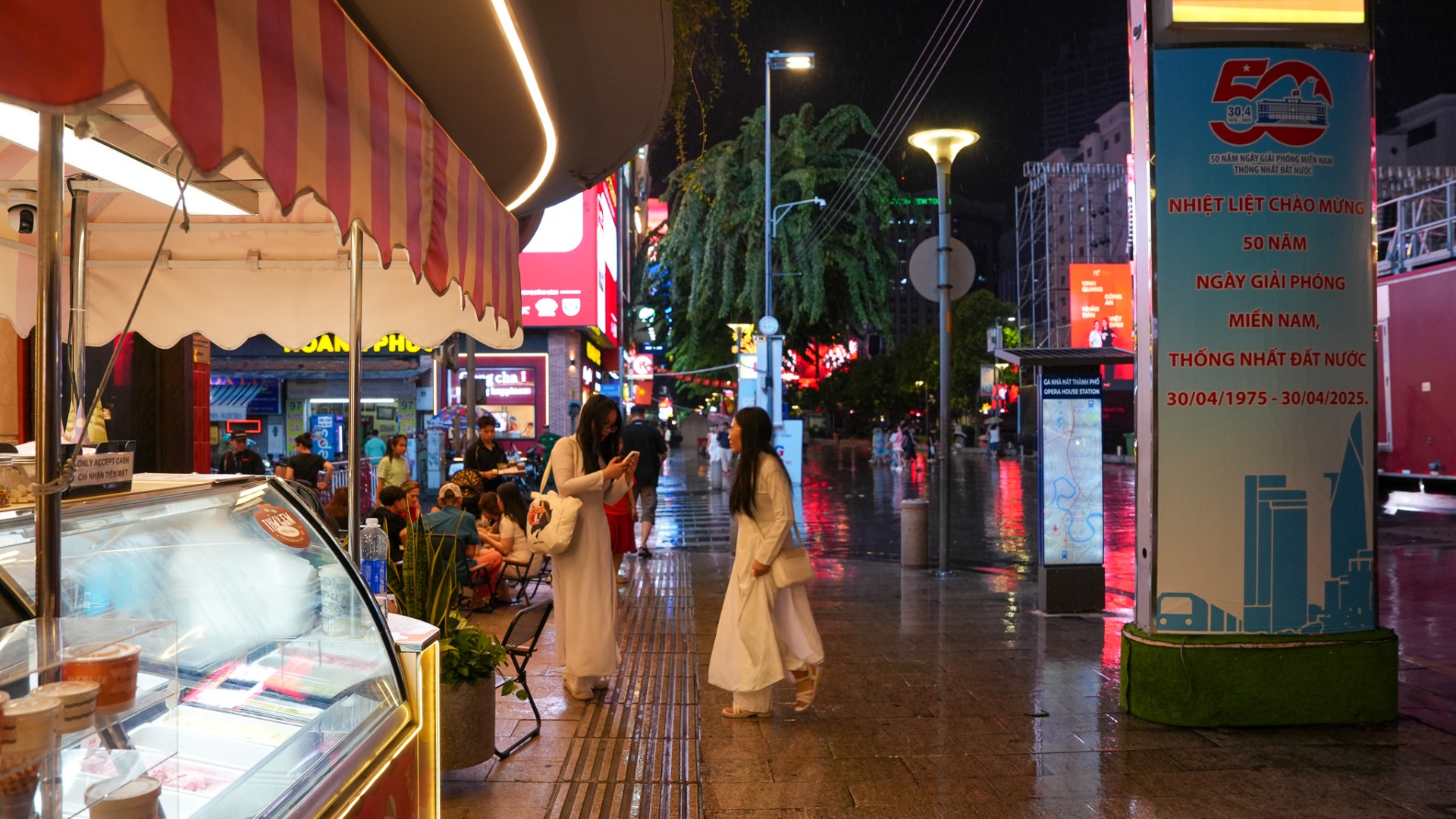 Two young women in white ao dai standing beside an ice-cream cart on Nguyen Hue Walking Street near the Opera House metro station, District 1, Saigon