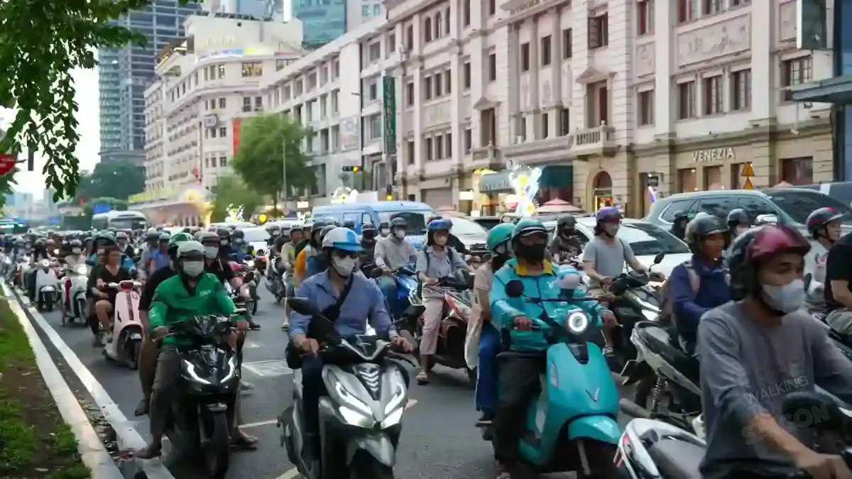 Head-on view of rush hour in District 1, Ho Chi Minh City — a dense wave of motorbike riders moving together past a French colonial façade in downtown Saigon