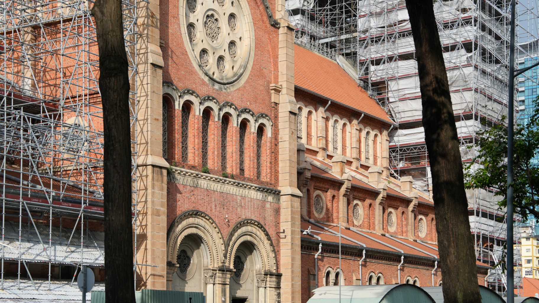 Front facade of Notre Dame Cathedral Saigon wrapped in restoration scaffolding, with the rose window and twin entrance arches still visible in District 1, Ho Chi Minh City