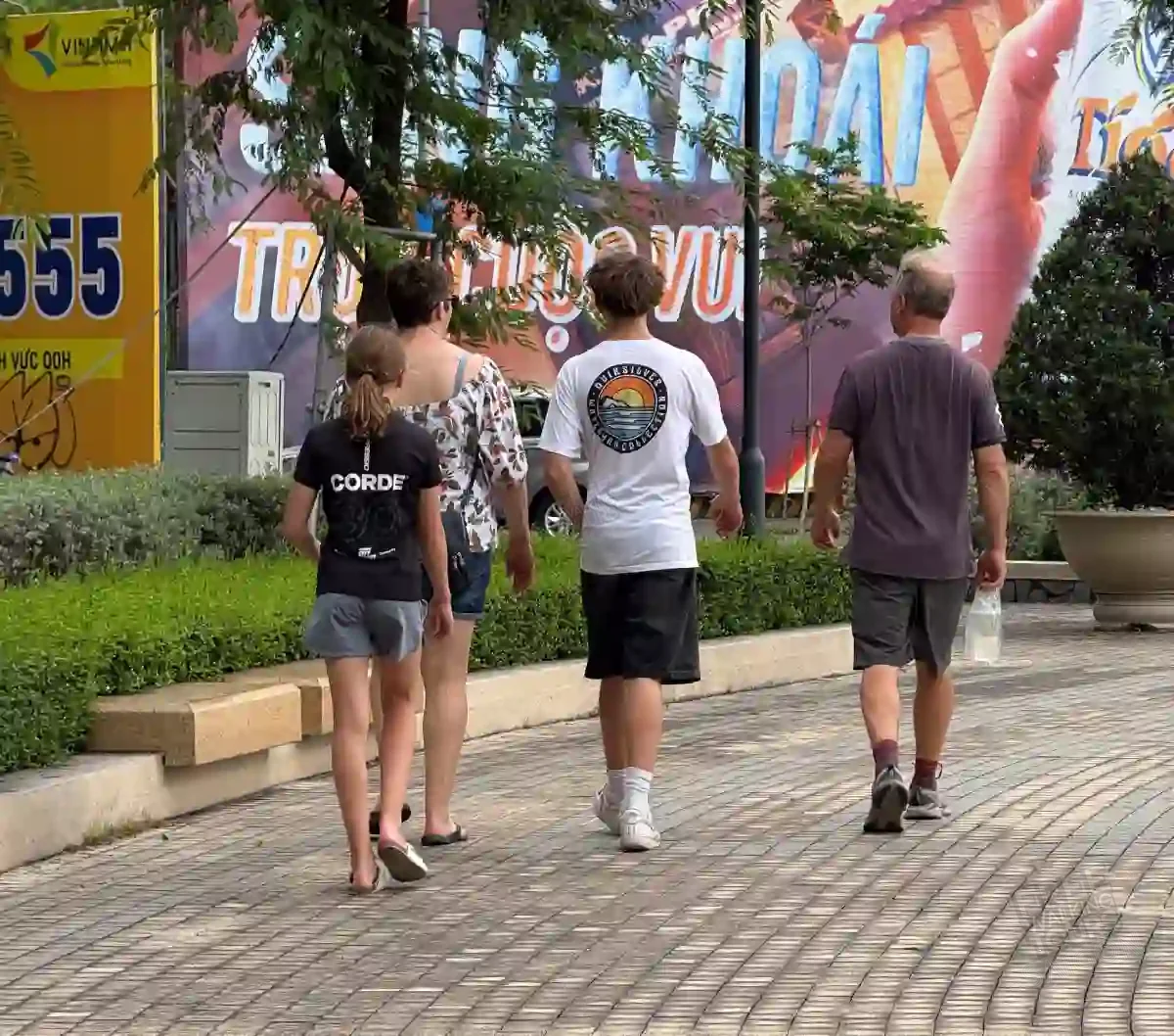 A tourist family walks calmly together along a tree-lined sidewalk in District 1, Saigon, after safely crossing a busy street