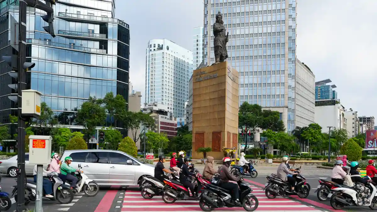 Motorbikes and cars crossing Me Linh Square roundabout beneath the Tran Hung Dao statue, surrounded by glass towers in District 1, Ho Chi Minh City