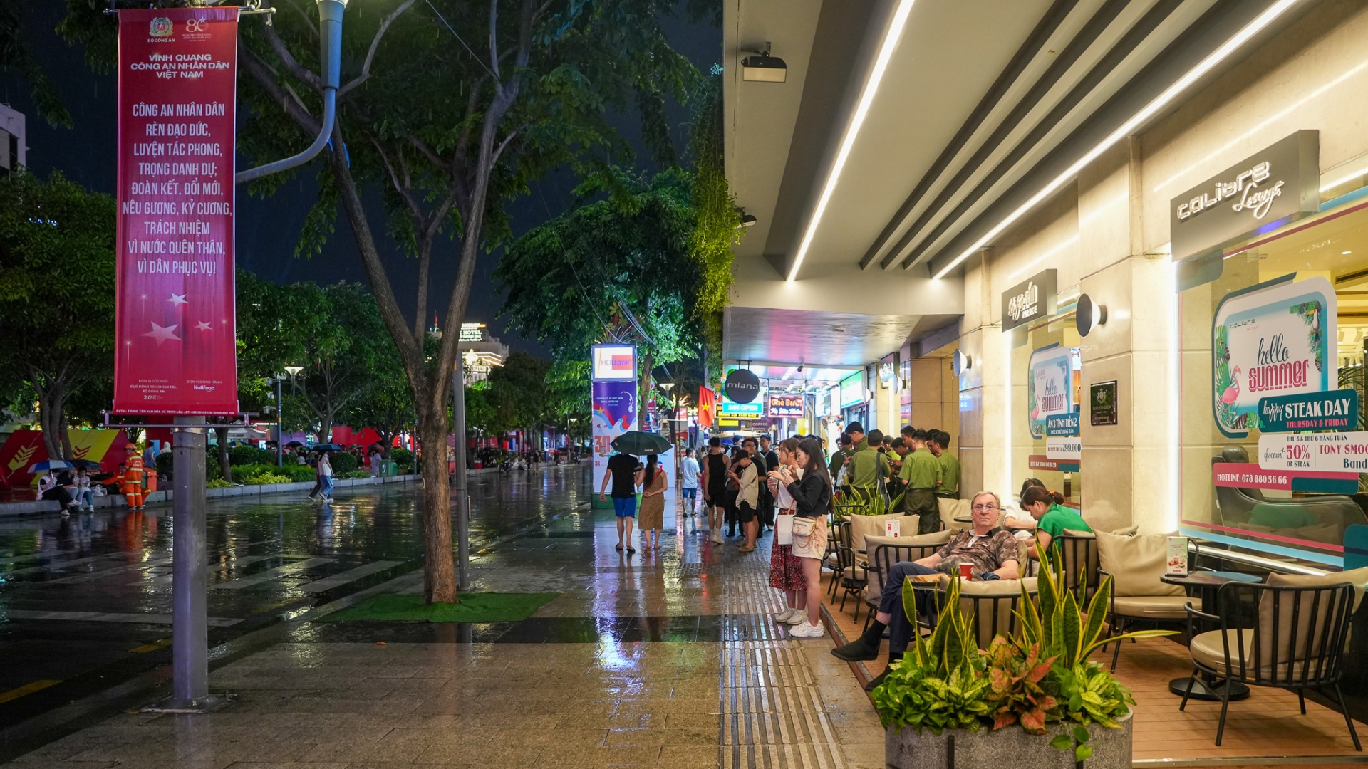 Locals and travellers relaxing at a covered sidewalk café along Nguyen Hue Walking Street on a rainy evening in District 1, Ho Chi Minh City