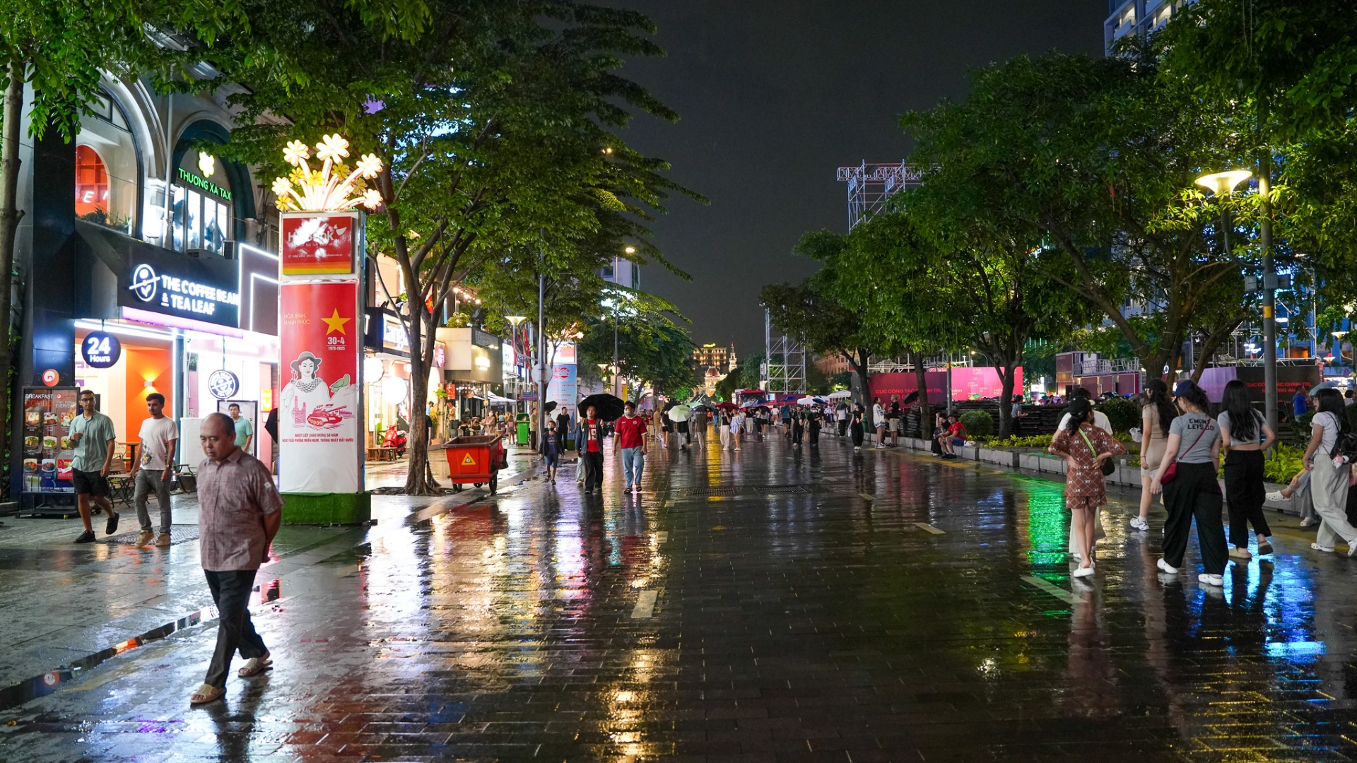 Nguyen Hue Walking Street at night after rain — tree-lined pedestrian boulevard with neon reflections on wet pavement and evening crowds in District 1, Ho Chi Minh City