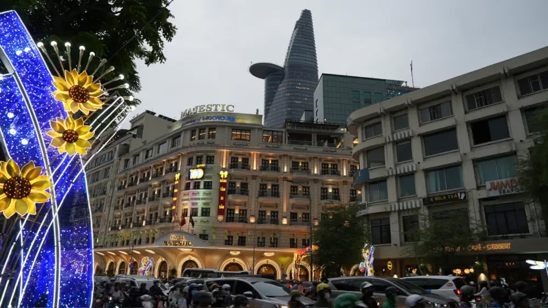 The colonial-era Hotel Majestic and the modern Bitexco Financial Tower seen together at dusk in District 1, Ho Chi Minh City, with festive sunflower lights and motorbike traffic in the foreground