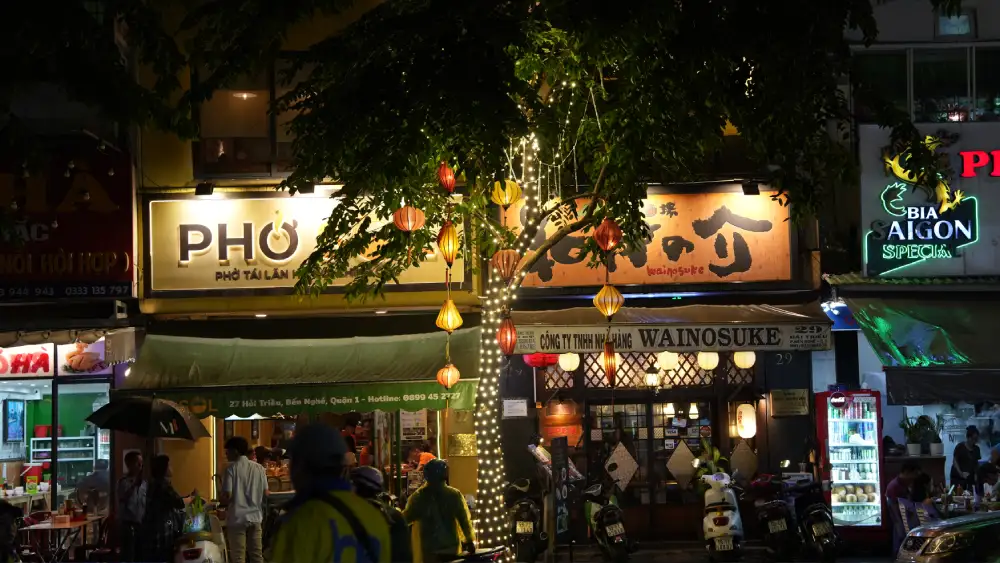 Side-by-side pho and Japanese restaurants on a District 1 street in Saigon at night, with silk lanterns strung between trees and locals stopping for a late-evening meal