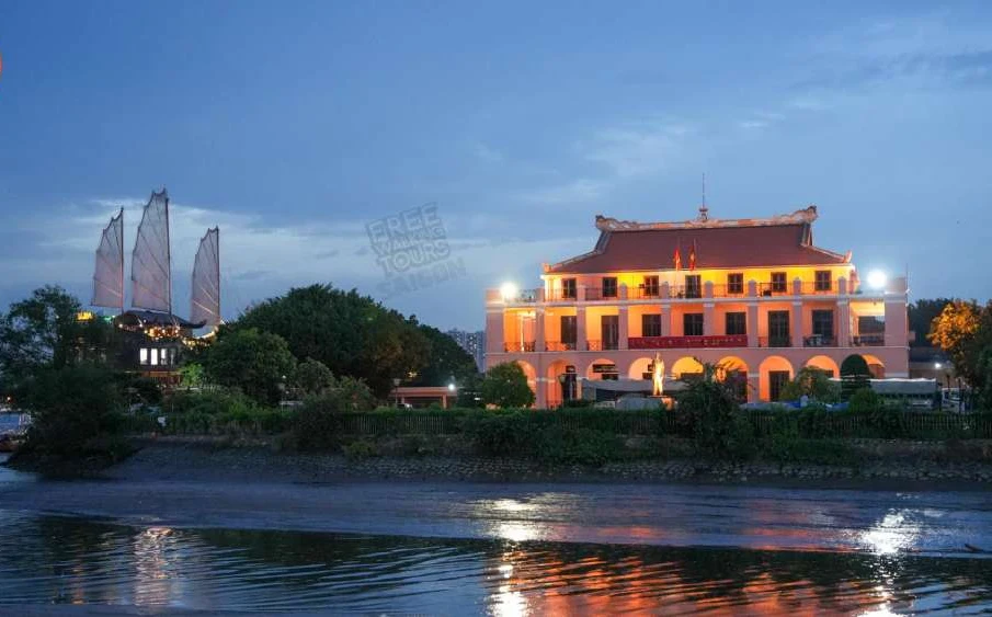 Nha Rong Harbour illuminated in warm light at night beside the Saigon River, with a traditional wooden junk boat with raised sails moored alongside